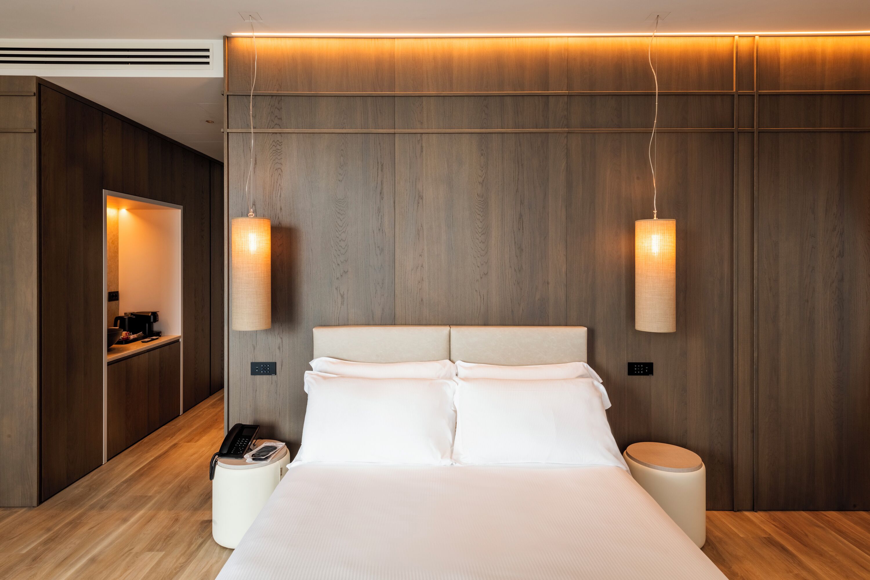 Una habitación de hotel moderna con un panel de pared de madera, una cama grande con ropa blanca, dos lámparas de noche, un teléfono junto a la cama y una mesita de noche con lámpara.