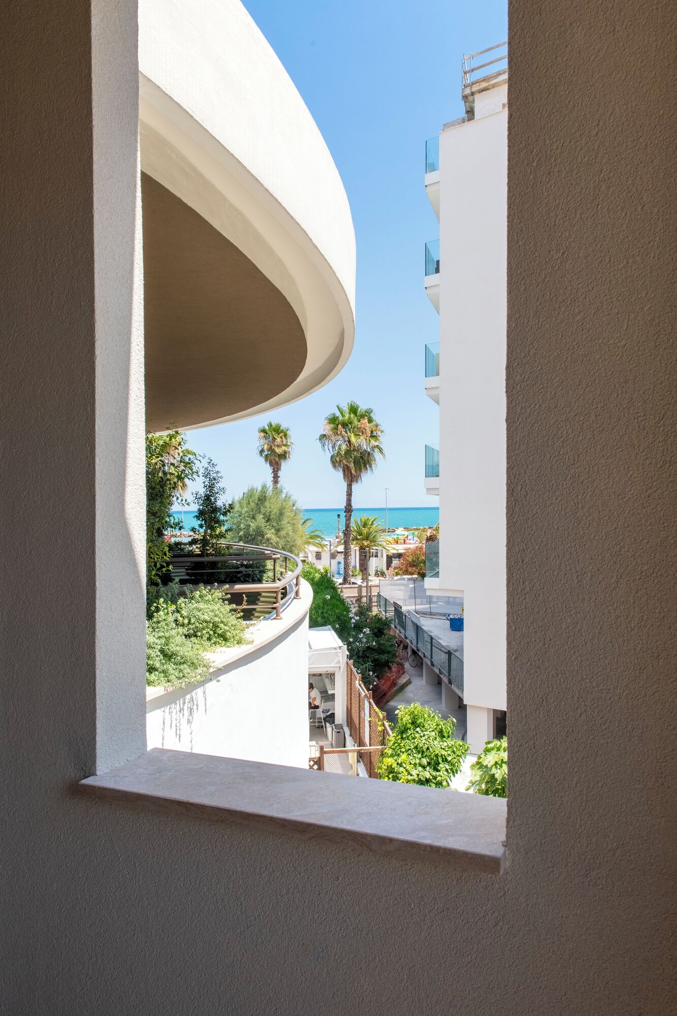 Un balcón con una estructura blanca curva, con vista a una zona costera con palmeras y un cielo azul despejado.