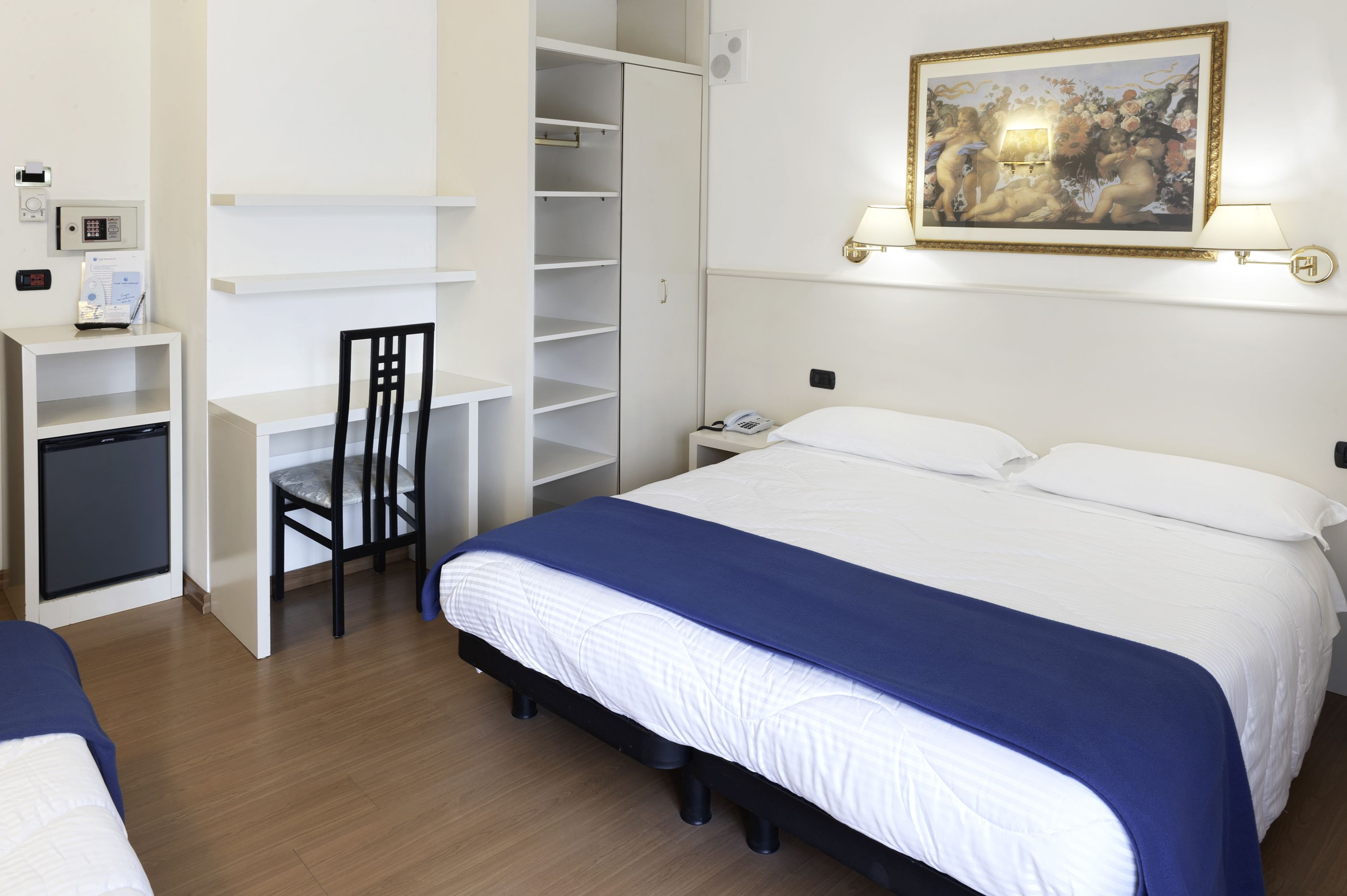 Une chambre d’hôtel comprenant un lit, un bureau avec une chaise, une armoire, une table de chevet avec une lampe et un tableau au mur.