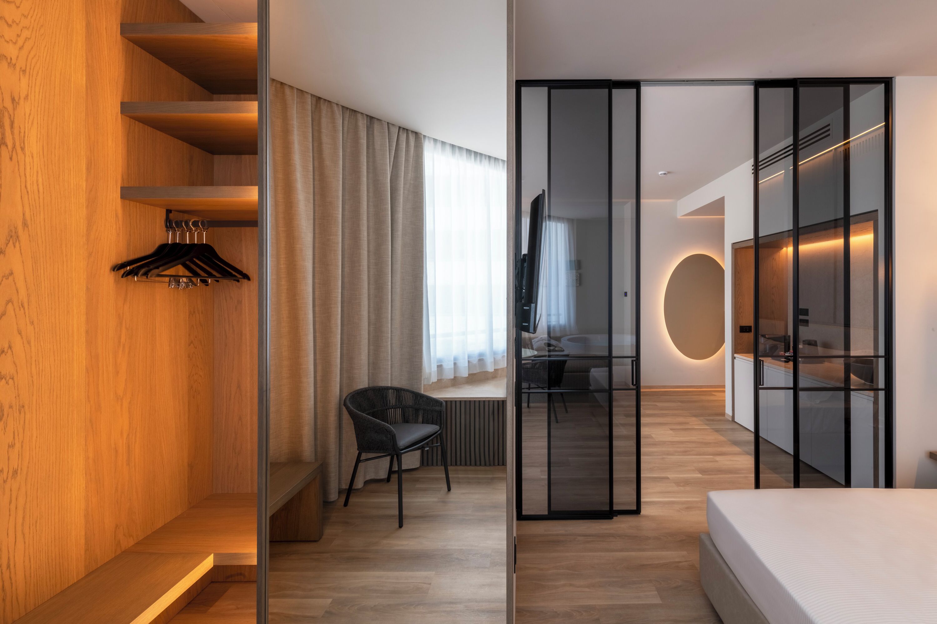 A modern hotel room with a wooden accent wall, a seating area with a chair, a bed, a desk, and a wardrobe with a mirror.