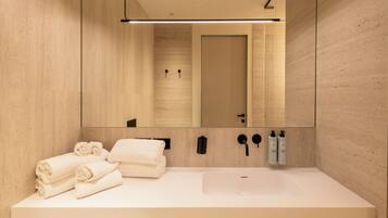 Une salle de bain moderne avec un grand miroir, un lavabo blanc, des serviettes soigneusement pliées et deux bouteilles de shampoing et d’après-shampoing.