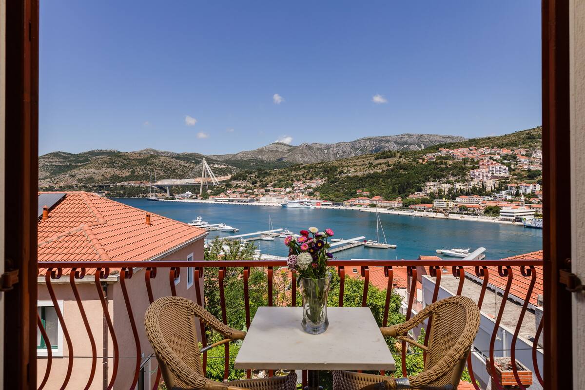 A balcony with wicker chairs, a table with a vase of flowers, and a view of a coastal town with boats and a bridge.