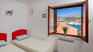 Een slaapkamer met twee bedden, een raam met uitzicht op een kustplaatsje en bergen, en een radiator onder het raam.