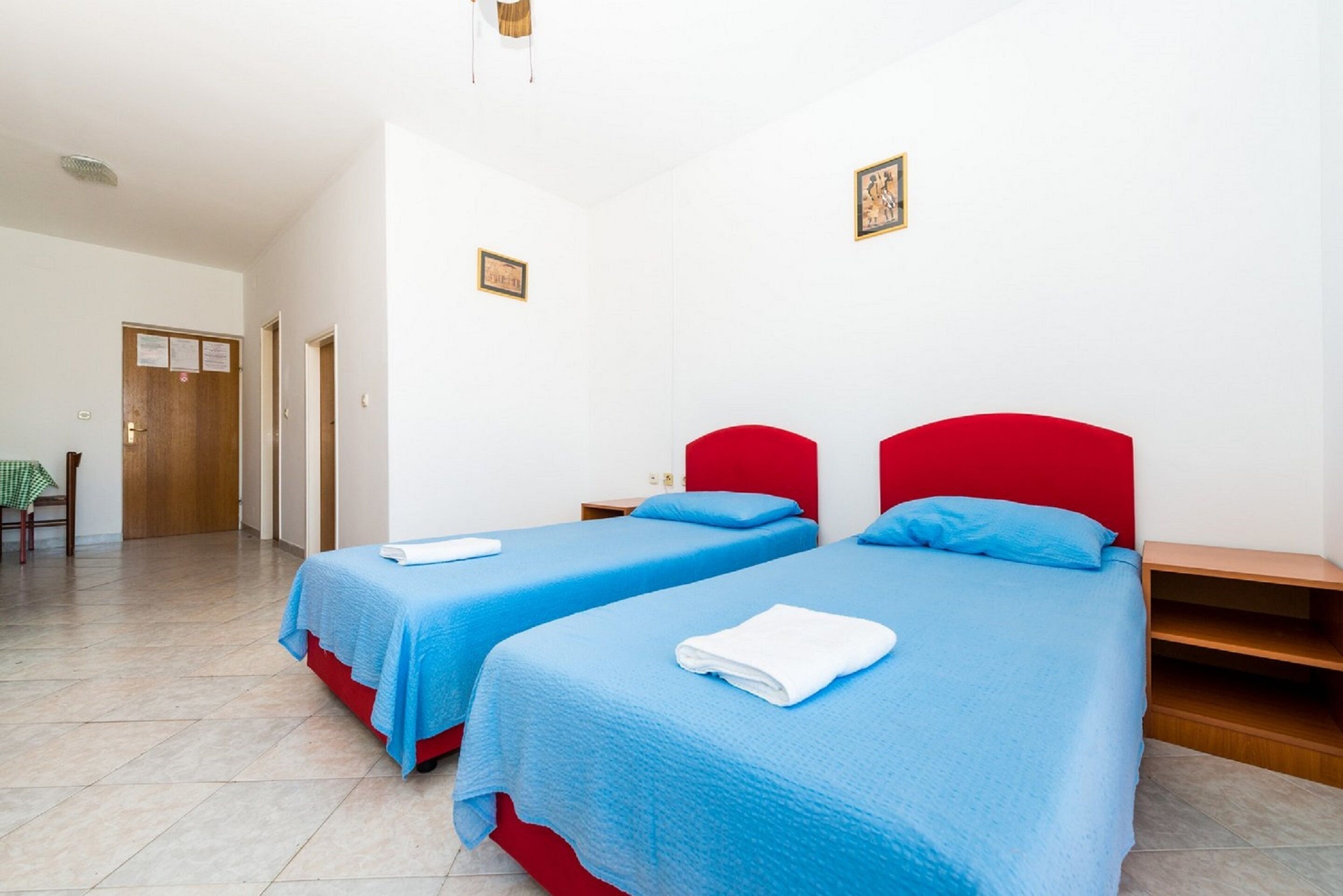 A hotel room with two beds, each with a blue bedspread and a red headboard. There is a wooden nightstand between the beds, and a small table with a green tablecloth in the corner.