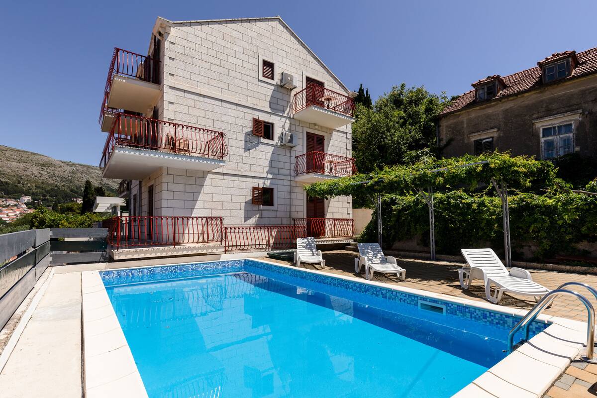 A two-story building with a swimming pool, white walls, balconies with red railings, and white lounge chairs.