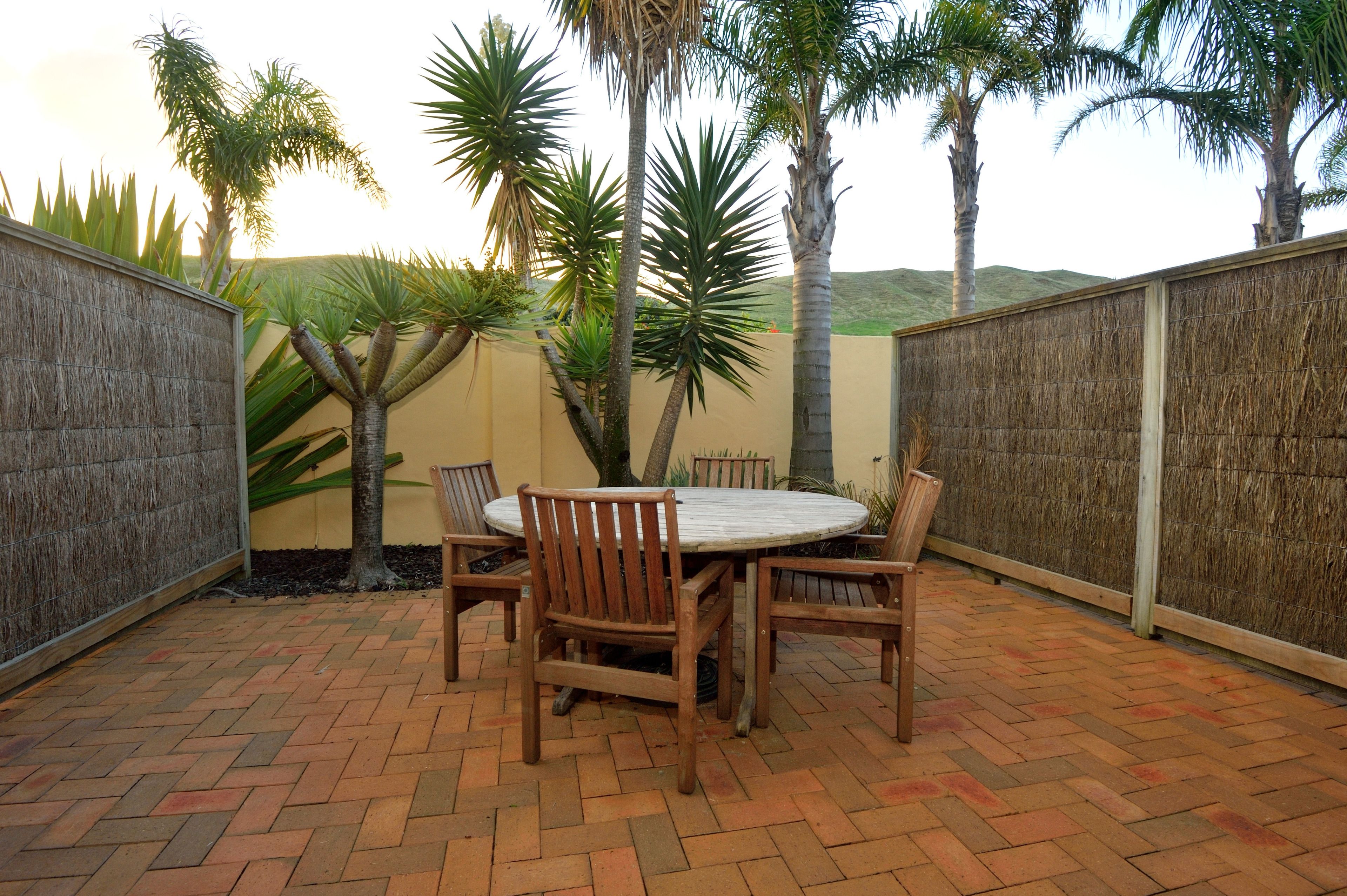 An outdoor patio with a round table and chairs, surrounded by palm trees and a brick floor.