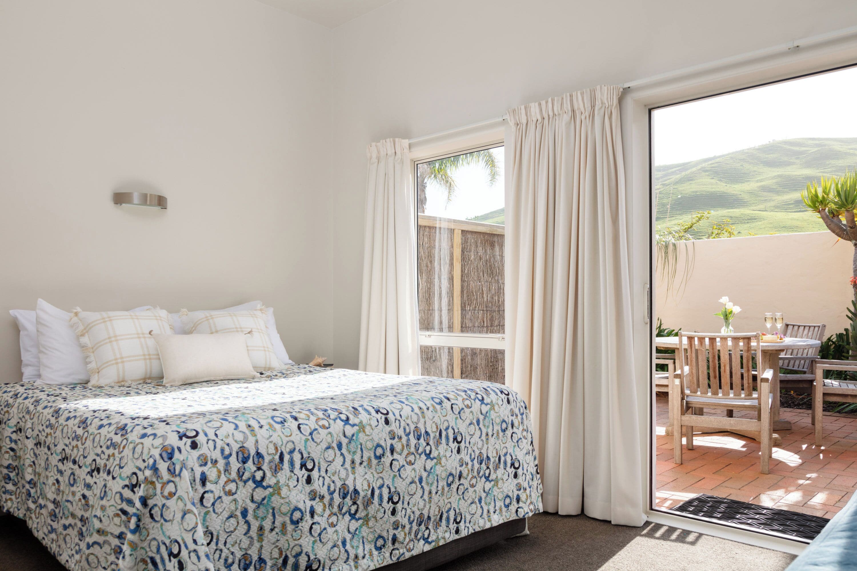 Un dormitorio con cama, ventana y vista a un patio con mobiliario exterior.