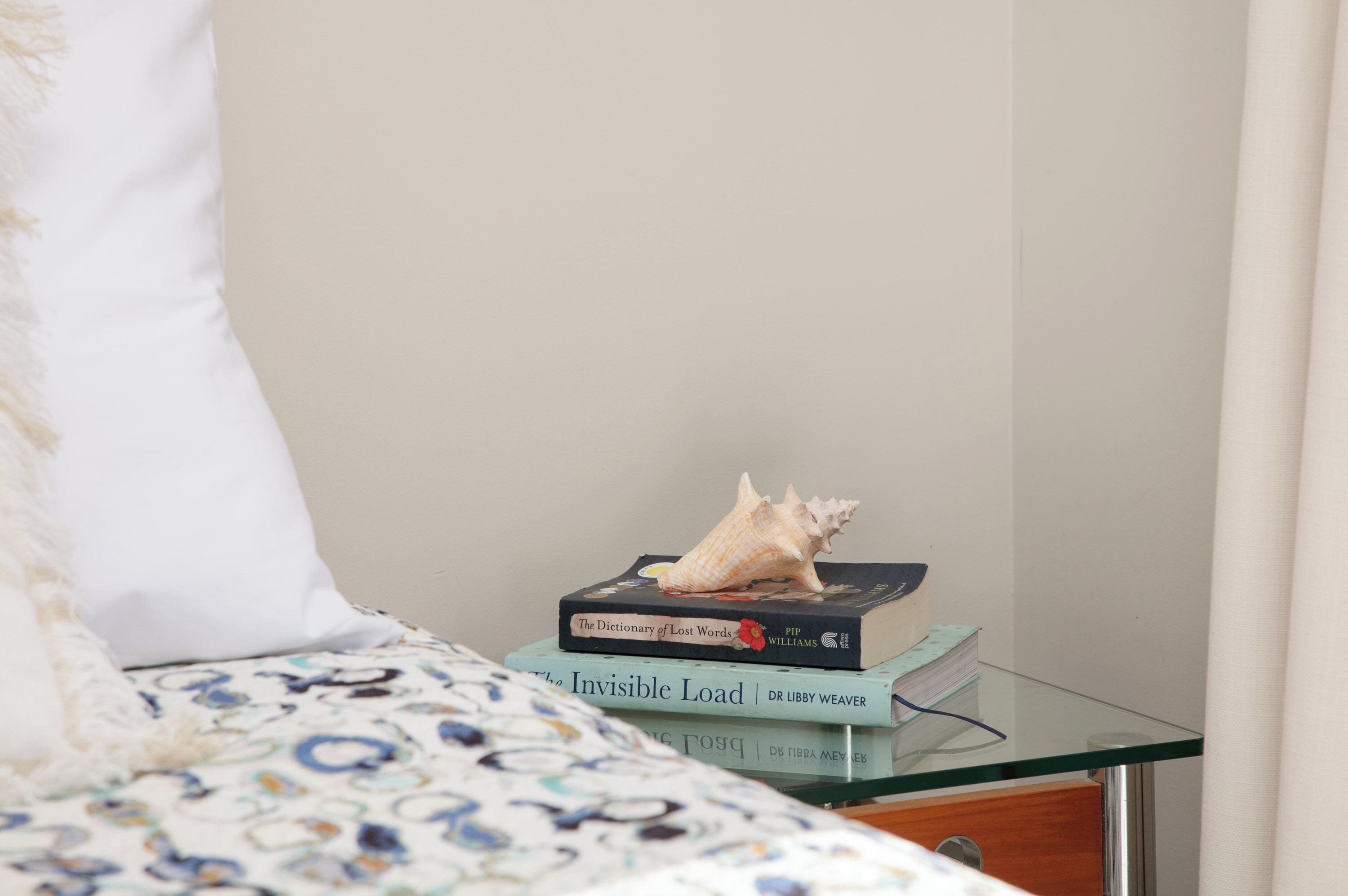 Una cama con un edredón estampado, una almohada blanca y una mesita de cristal con dos libros y una concha.