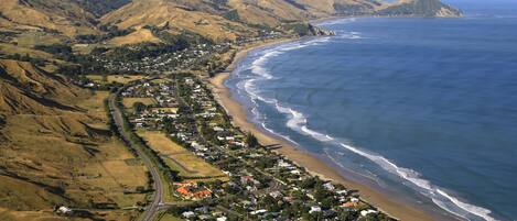 An aerial view of a coastal town with a beach, hilly terrain, and a road running alongside the coastline.