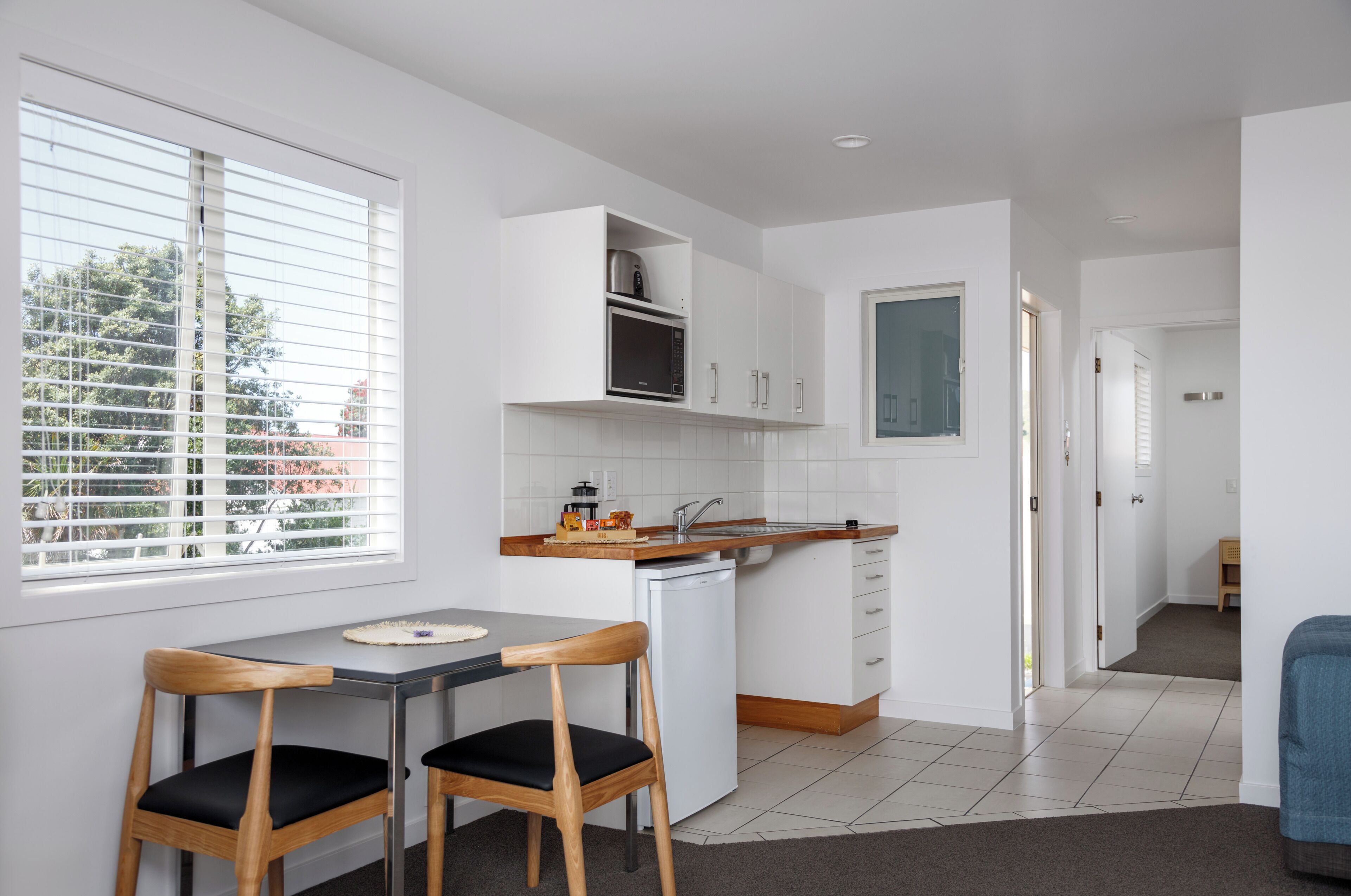 A compact kitchen with white cabinets, a built-in oven, and a dishwasher. There is a dining table with two chairs and a window with blinds.