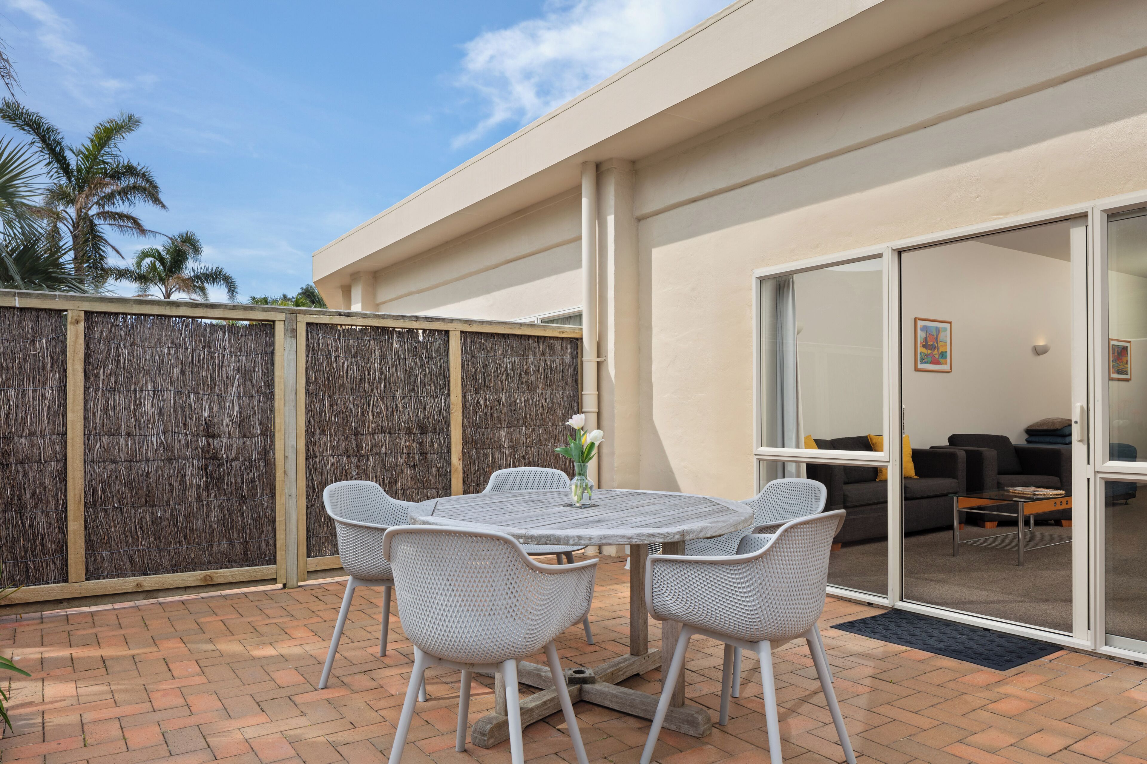 A patio with a round table and chairs, a woven fence, and a sliding glass door leading to an interior space with a sofa and a coffee table.