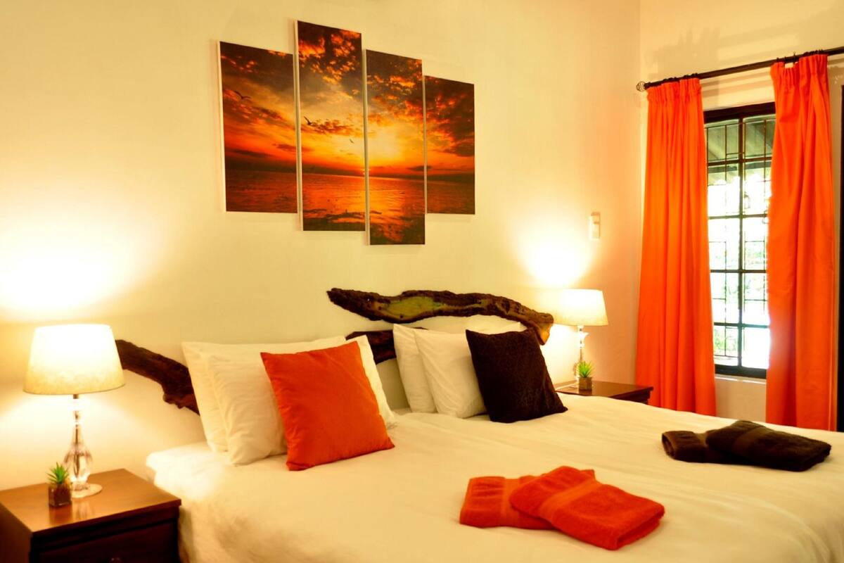 A hotel room with a large bed, two bedside tables, a nightstand, a window with orange curtains, and a sunset painting on the wall.