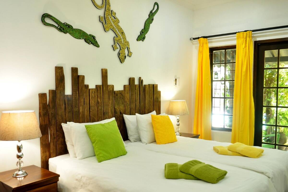 A hotel room with a wooden headboard, a large bed with white bedding and colorful pillows, two bedside lamps, a nightstand, and a window with yellow curtains.