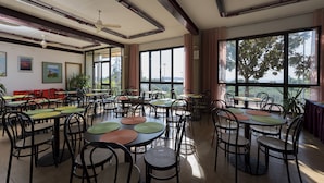 Un comedor amplio e interior con varias mesas redondas y sillas, ventanas grandes con vistas a árboles y ventiladores de techo.