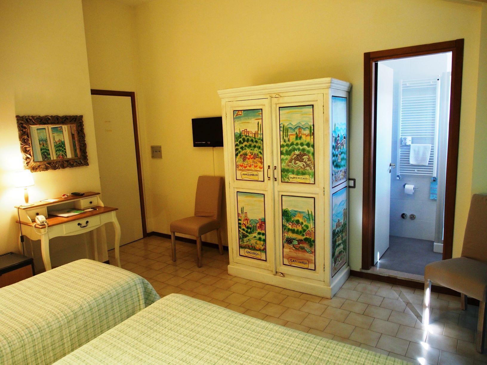 Ein Hotelzimmer mit zwei Betten, einem Schreibtisch mit Telefon, einem Sessel, einem Schrank mit verzierten Türen und einer Tür zu einem Badezimmer.