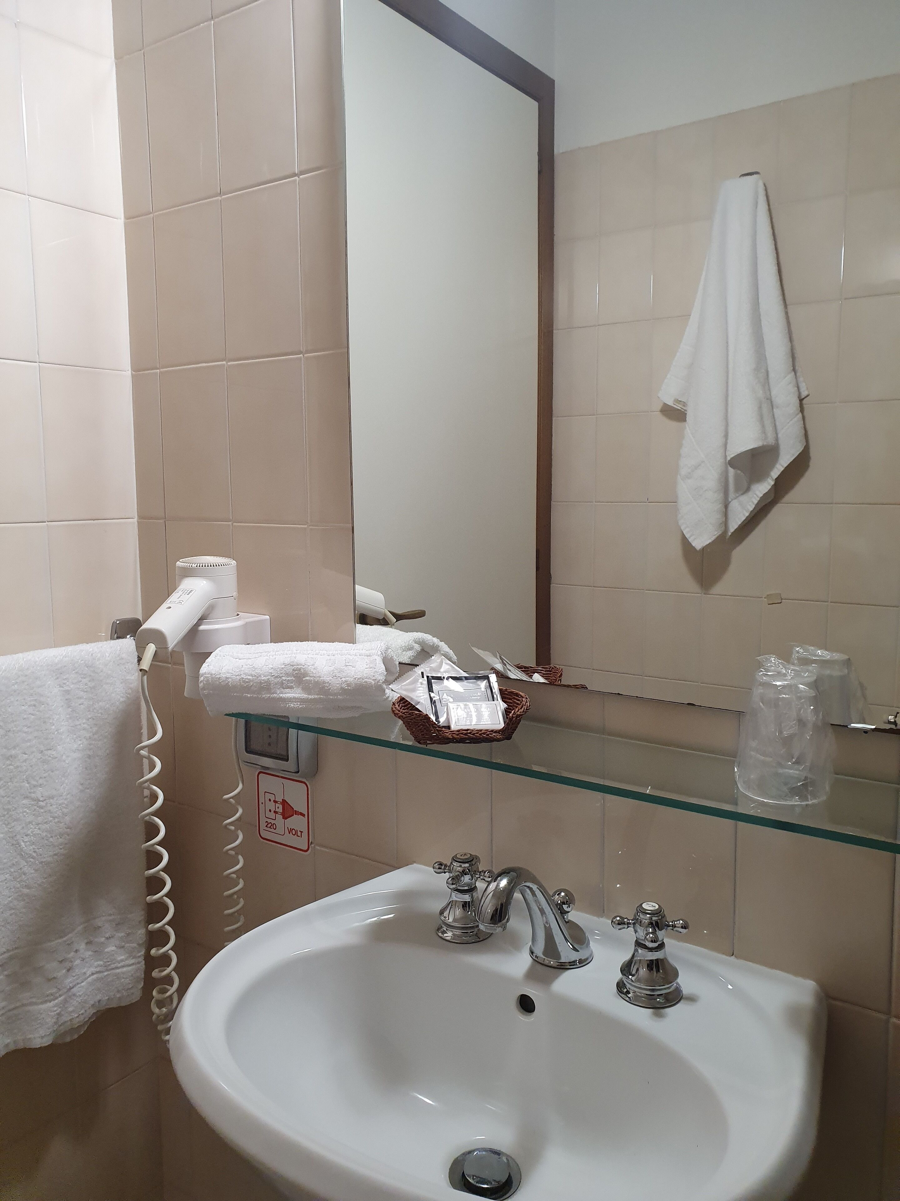 A bathroom with a white sink, two faucets, a mirror, a towel rack, and a glass shelf with toiletries.