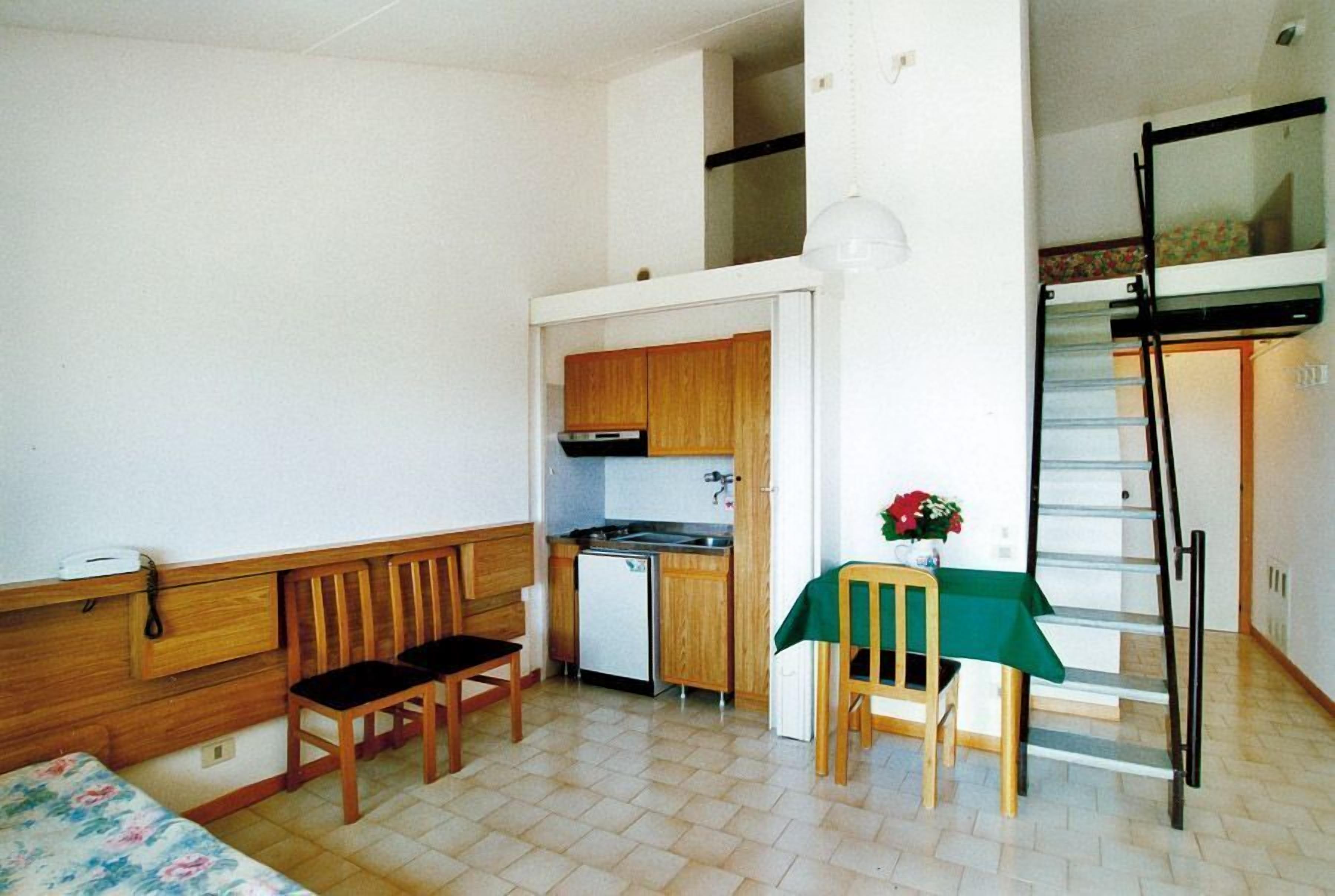 A compact room with a kitchenette, a dining area with a table and chairs, a bed, and a staircase.