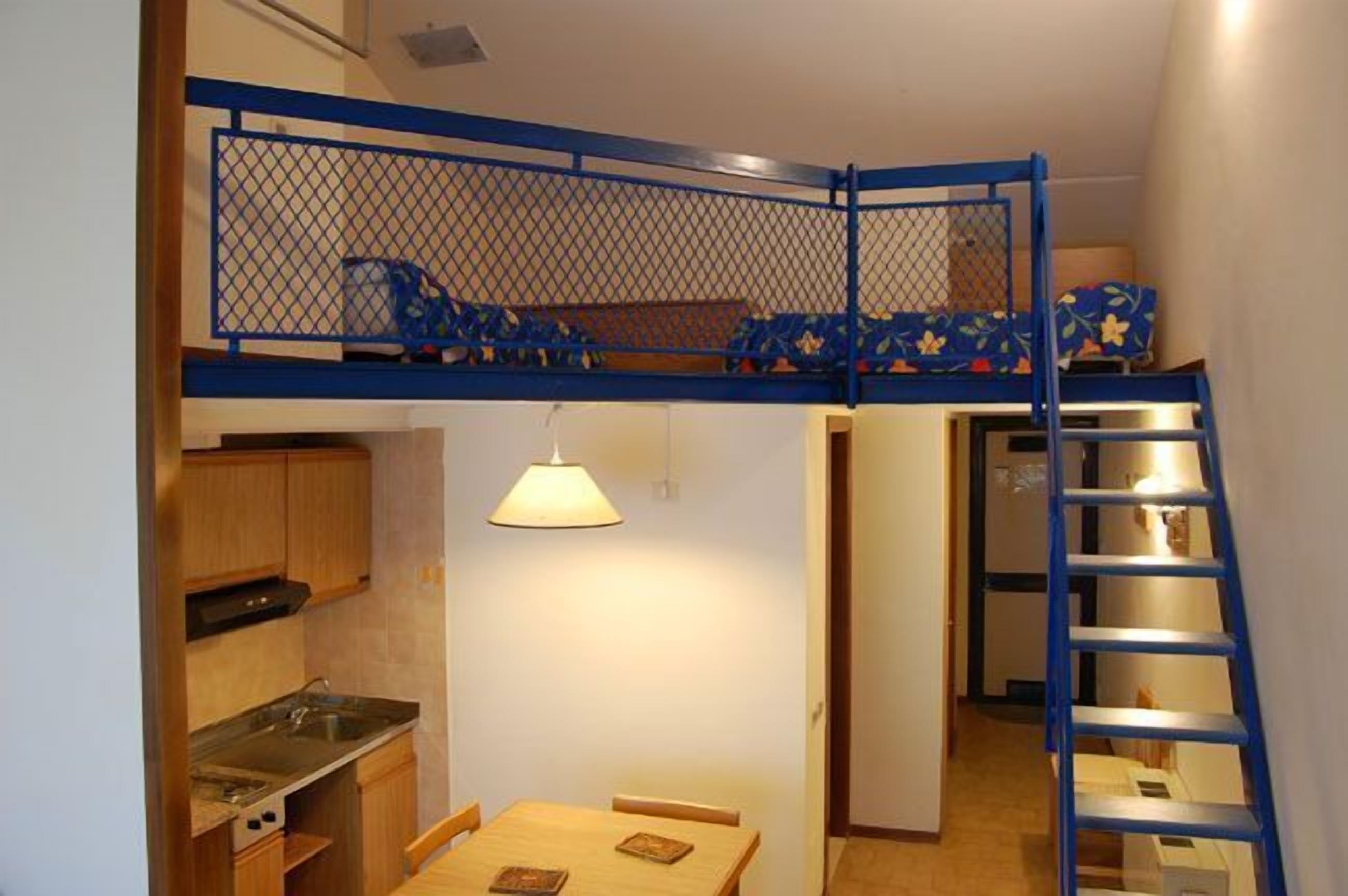 Ein kleines Zimmer mit Etagenbett, einer kompakten Küchenzeile, einem Esstisch und einer Treppe.