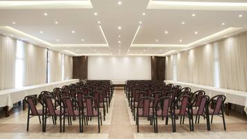 A conference room with rows of chairs, a long table, and a plain wall with curtains.