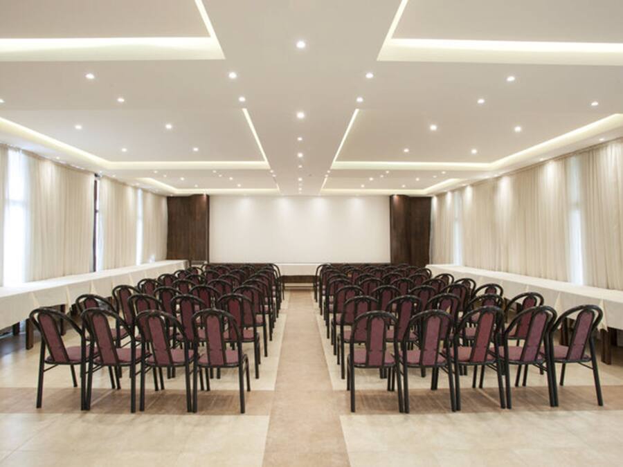A conference room with rows of chairs, a long table, and a plain wall with curtains.