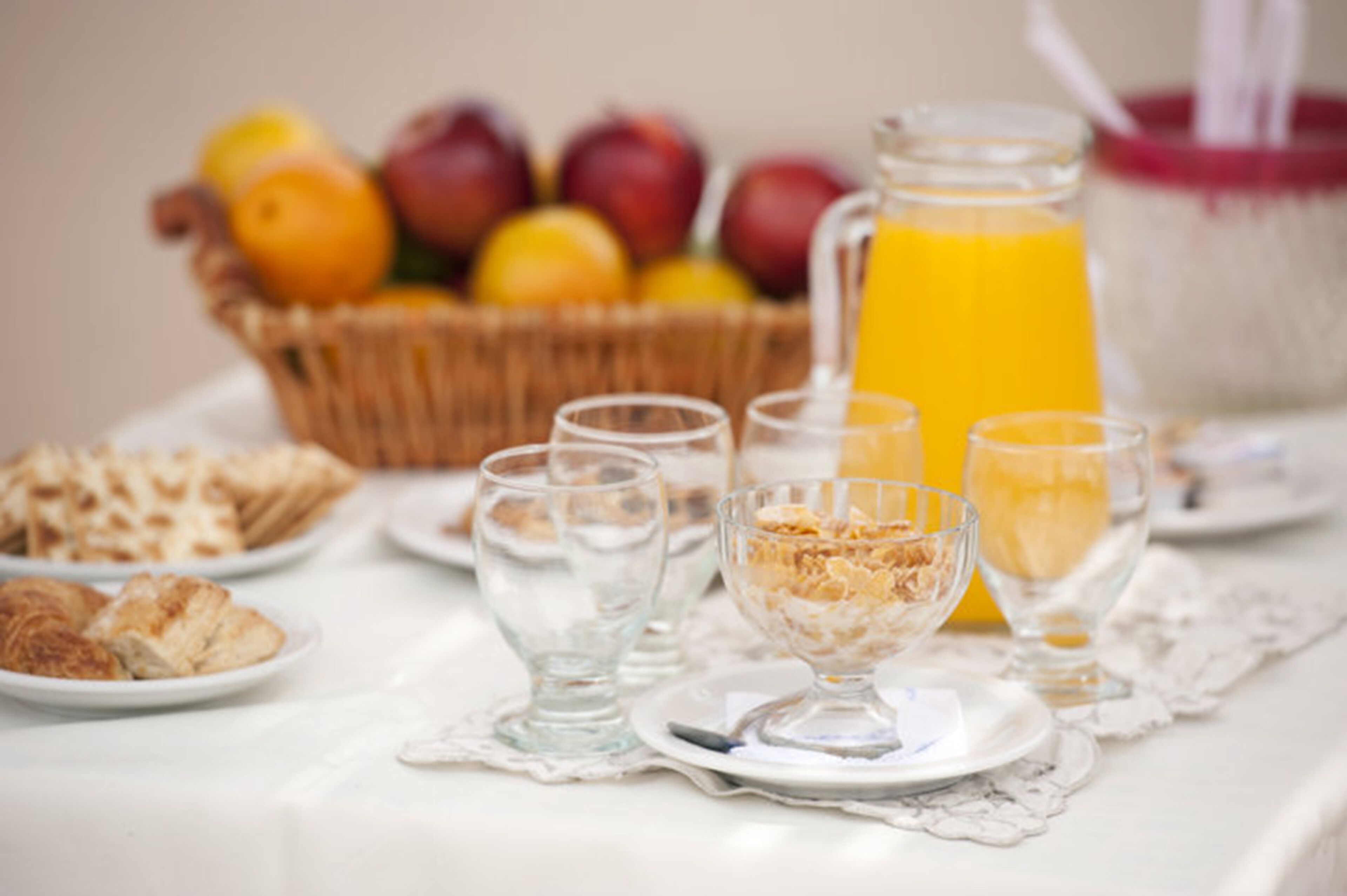 Een ontbijtsetting met een fruitmand, een karaf sinaasappelsap en diverse ontbijtspullen op een tafel.