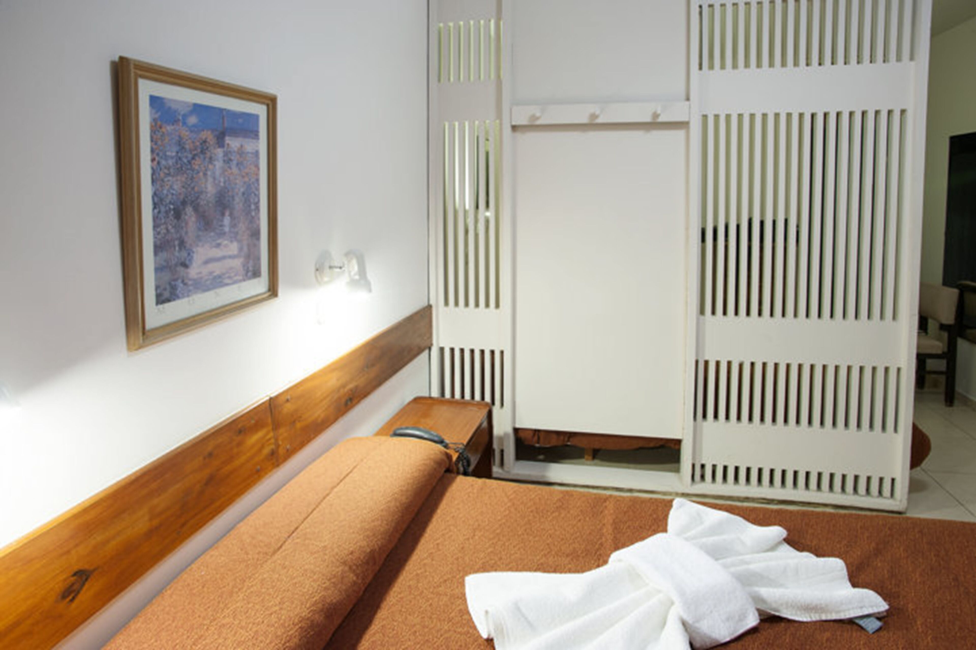 Une chambre avec un lit, une tête de lit en bois, une cloison blanche, un tableau encadré au mur et une serviette blanche sur le lit.