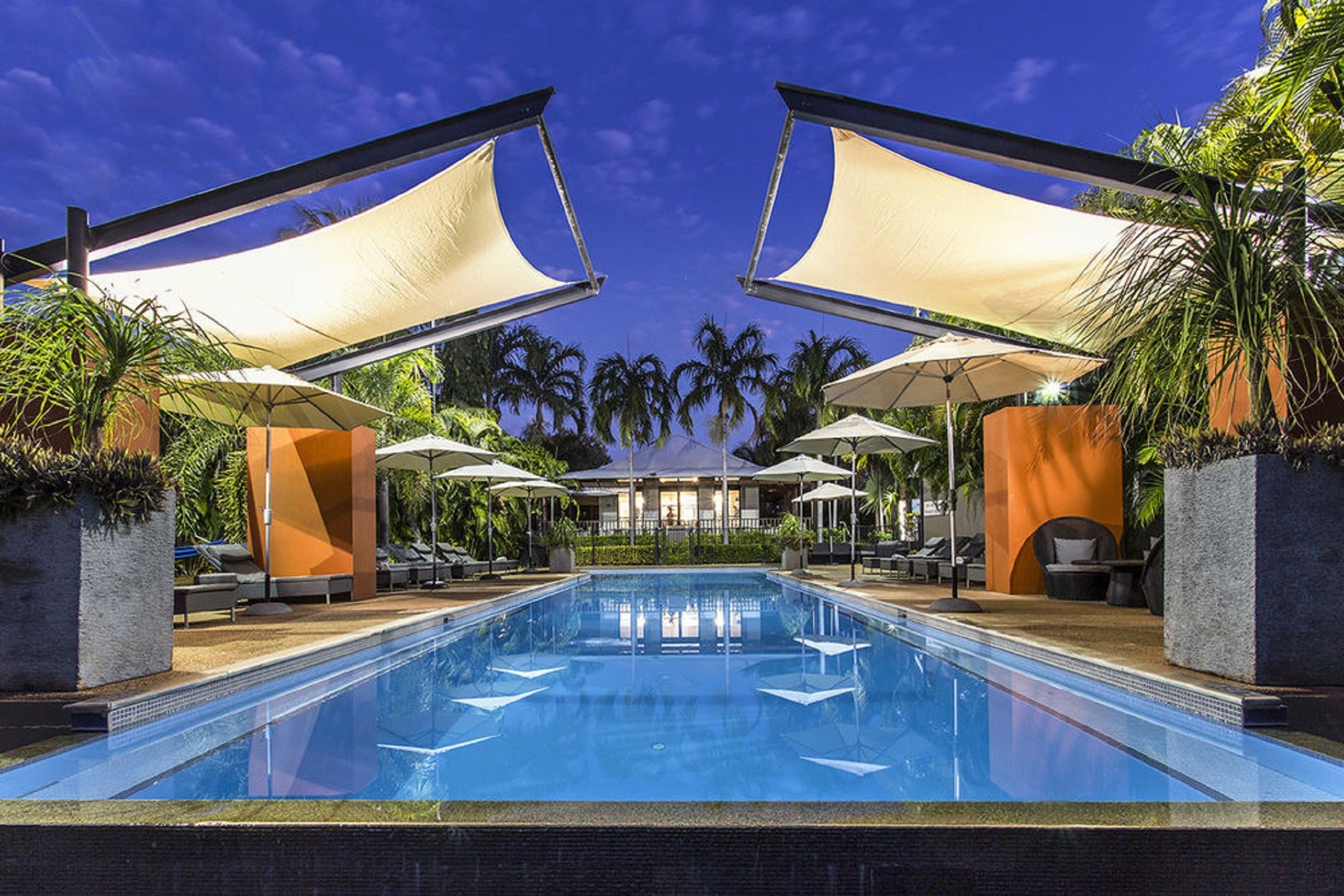 A swimming pool with lounge chairs and umbrellas, surrounded by palm trees and a building in the background.