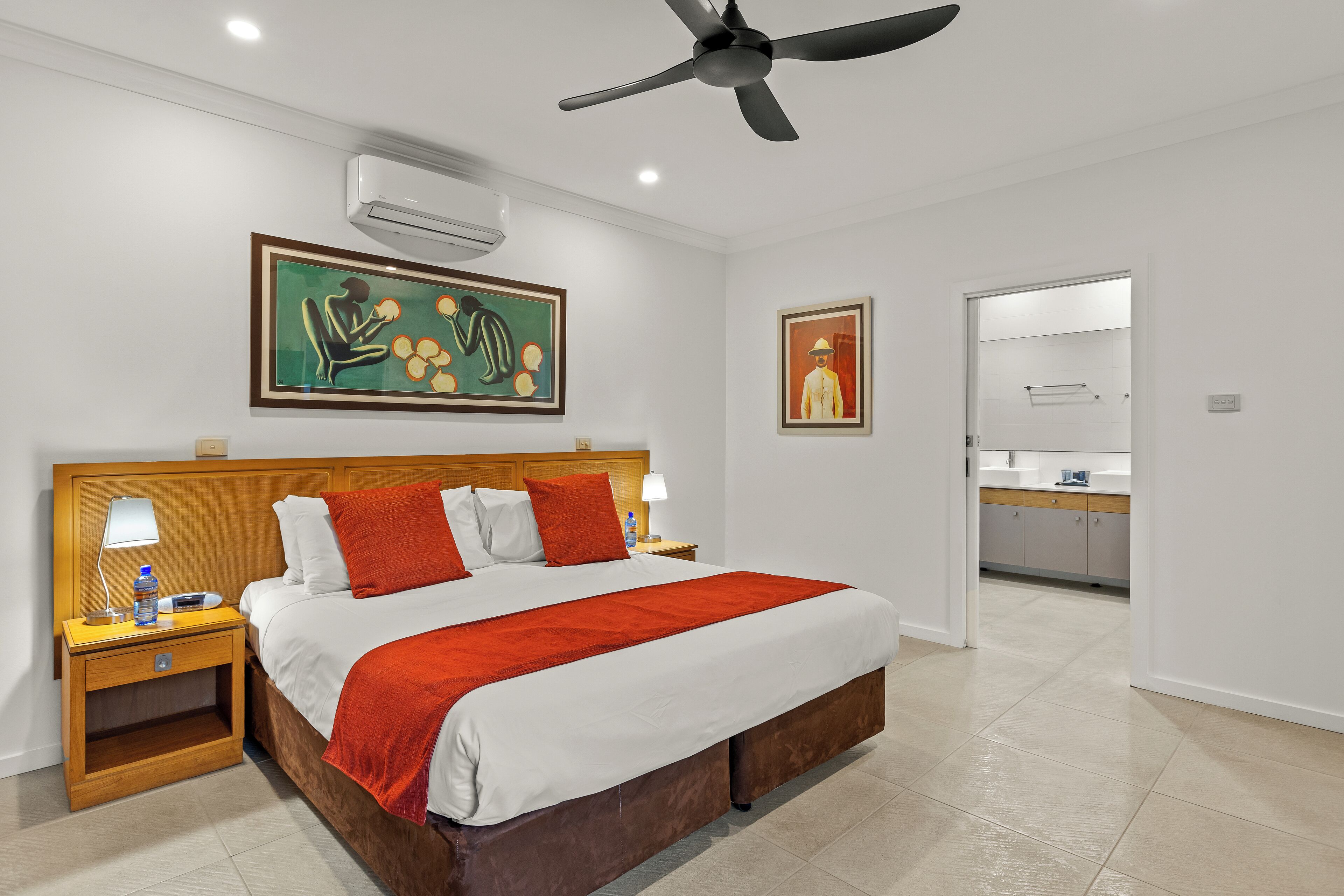 Habitación de hotel con una cama grande, un cabecero de madera, un ventilador de techo, aire acondicionado, una mesita de noche con una lámpara y una botella, y un cuadro en la pared.