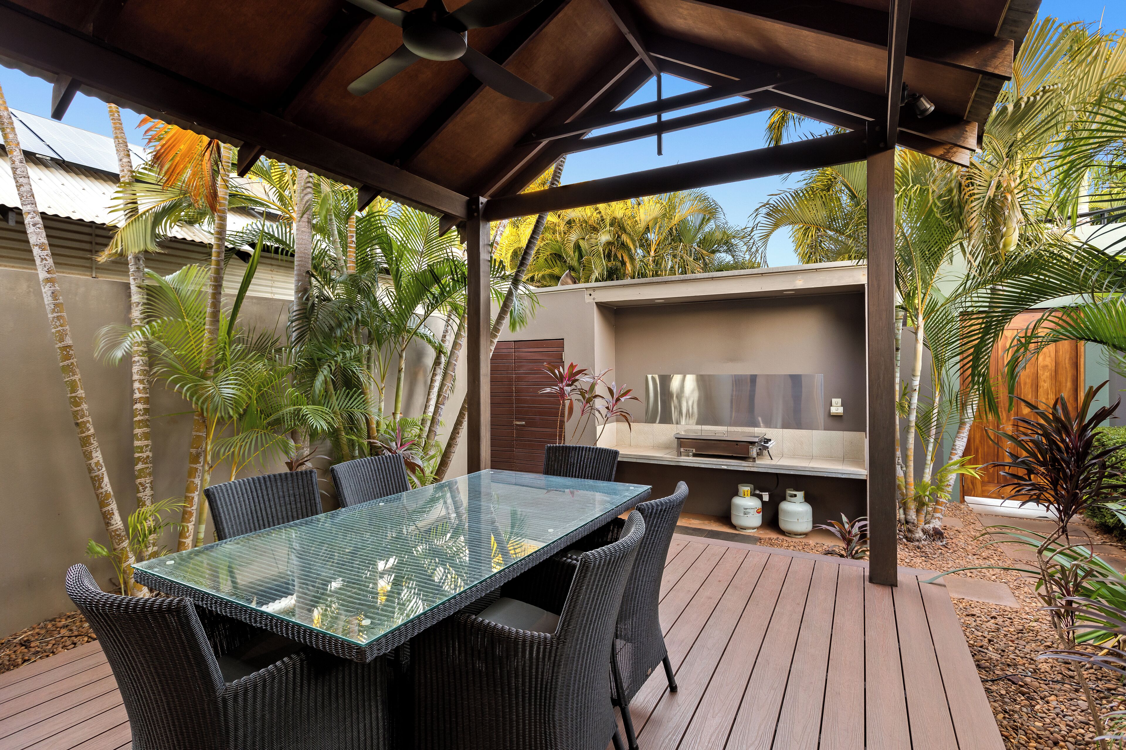 An outdoor patio with a glass-top table and wicker chairs, surrounded by palm trees and a barbecue area.