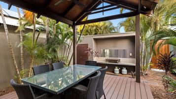 An outdoor patio with a glass-top table and wicker chairs, surrounded by palm trees and a barbecue area.