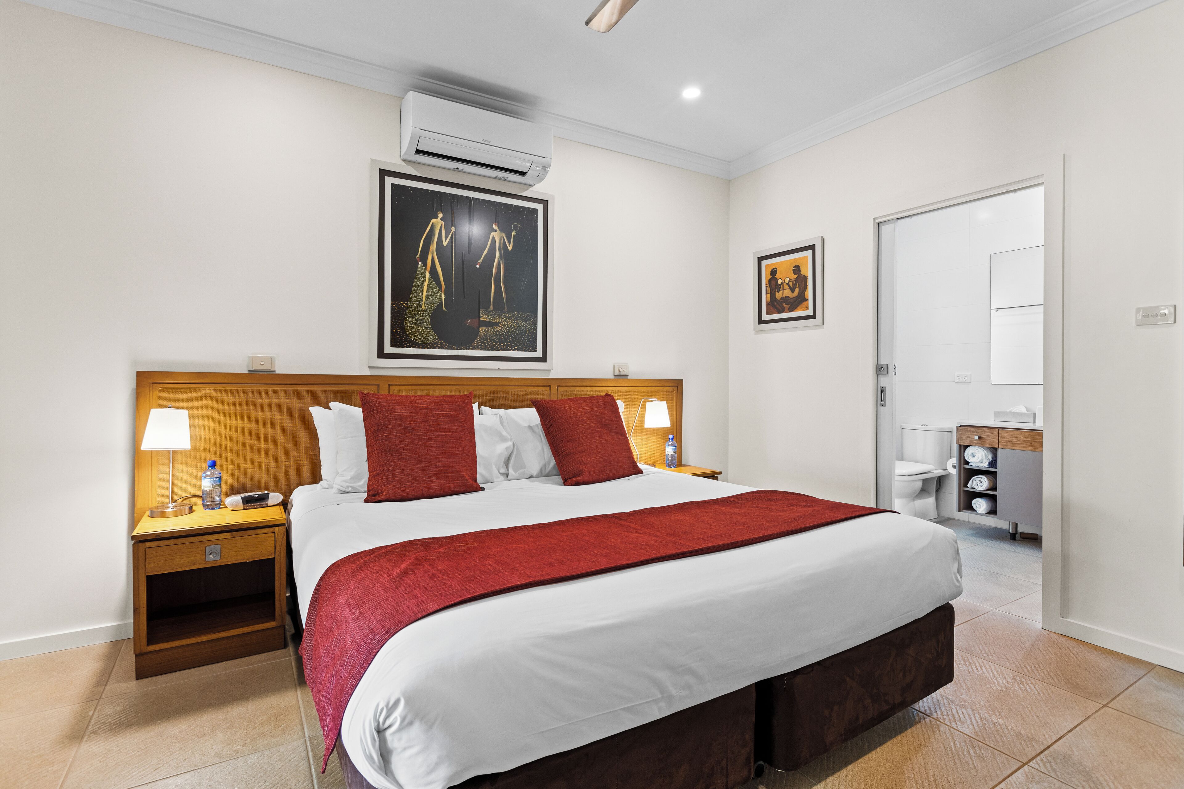 Habitación de hotel con una cama grande, un cabecero de madera, una mesita de noche con lámpara, aire acondicionado y un cuadro en la pared.