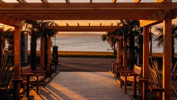 Une pergola en bois avec des bancs, donnant sur une plage au coucher du soleil.