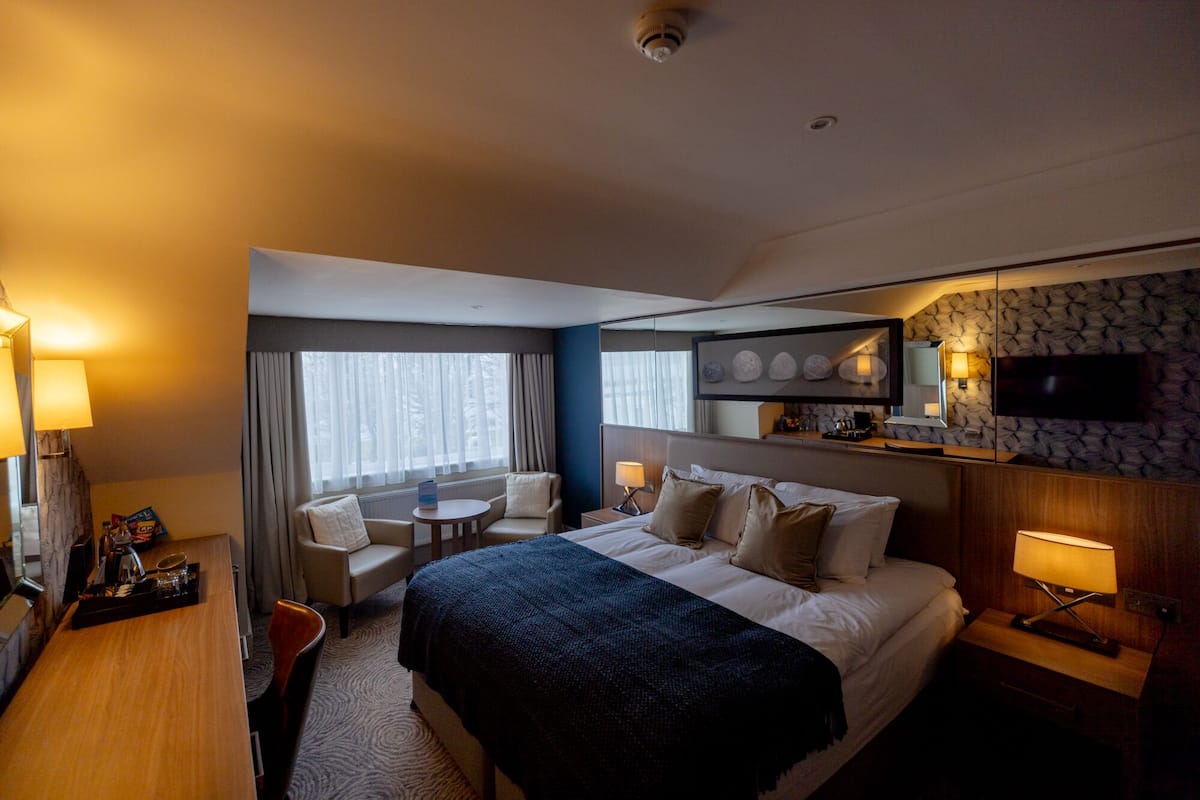 A hotel room with a bed, bedside tables, a desk, a chair, a small table, a sofa, a window with curtains, a mirror, and a television.