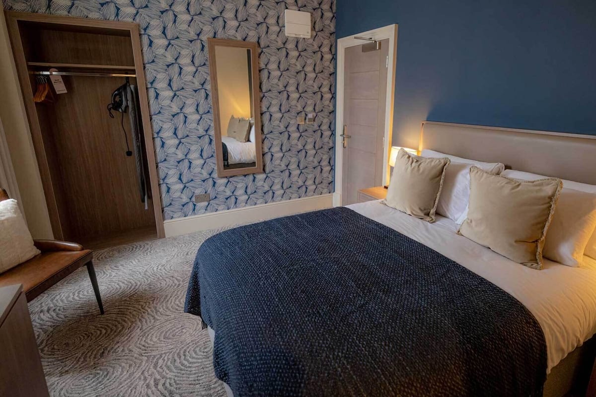 A hotel room with a bed, a bedside table, a nightstand, a wardrobe, a mirror, and a door.