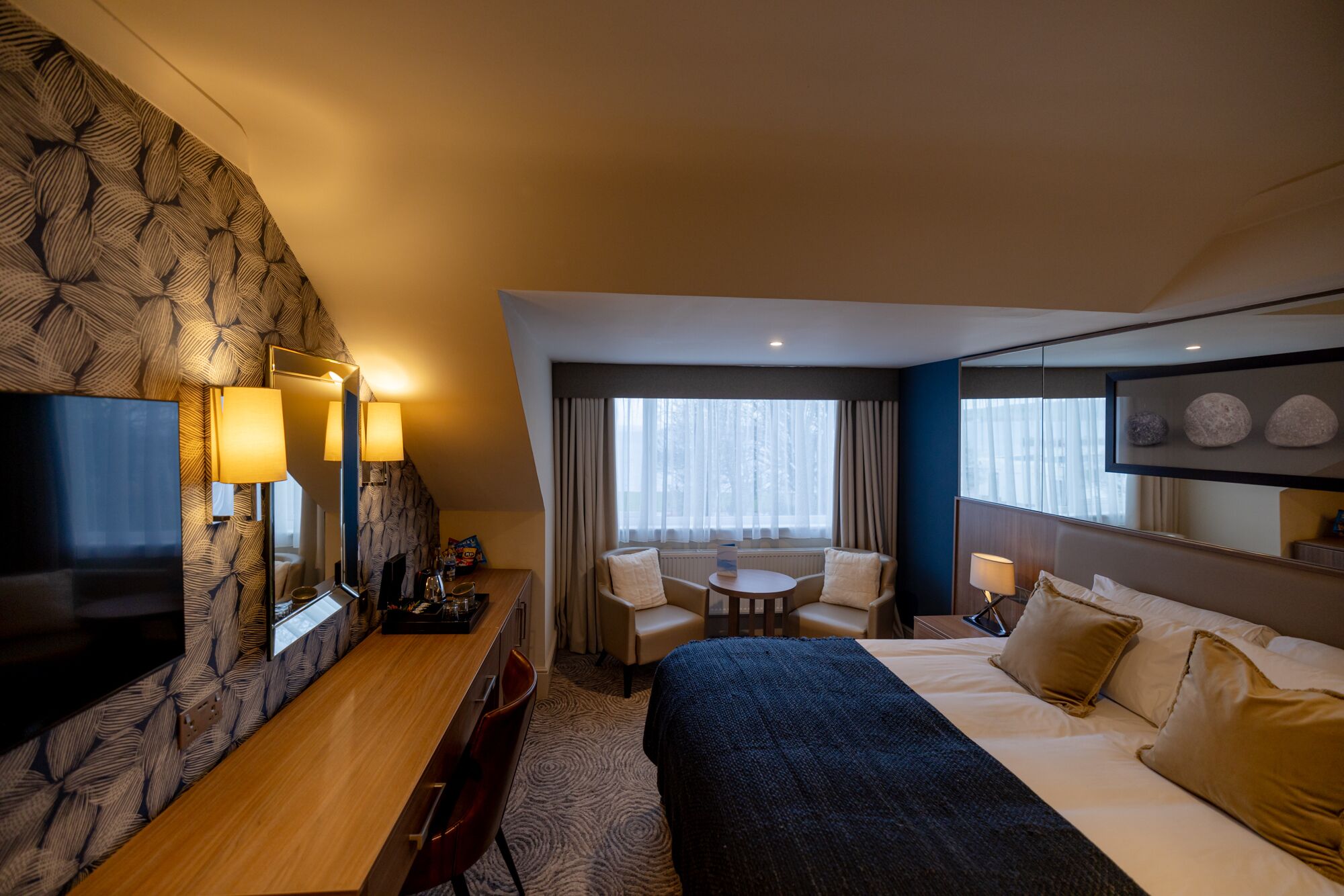 Una camera d'albergo con un letto, una scrivania con televisore, un angolo salotto con poltrone e un'ampia finestra con tende.