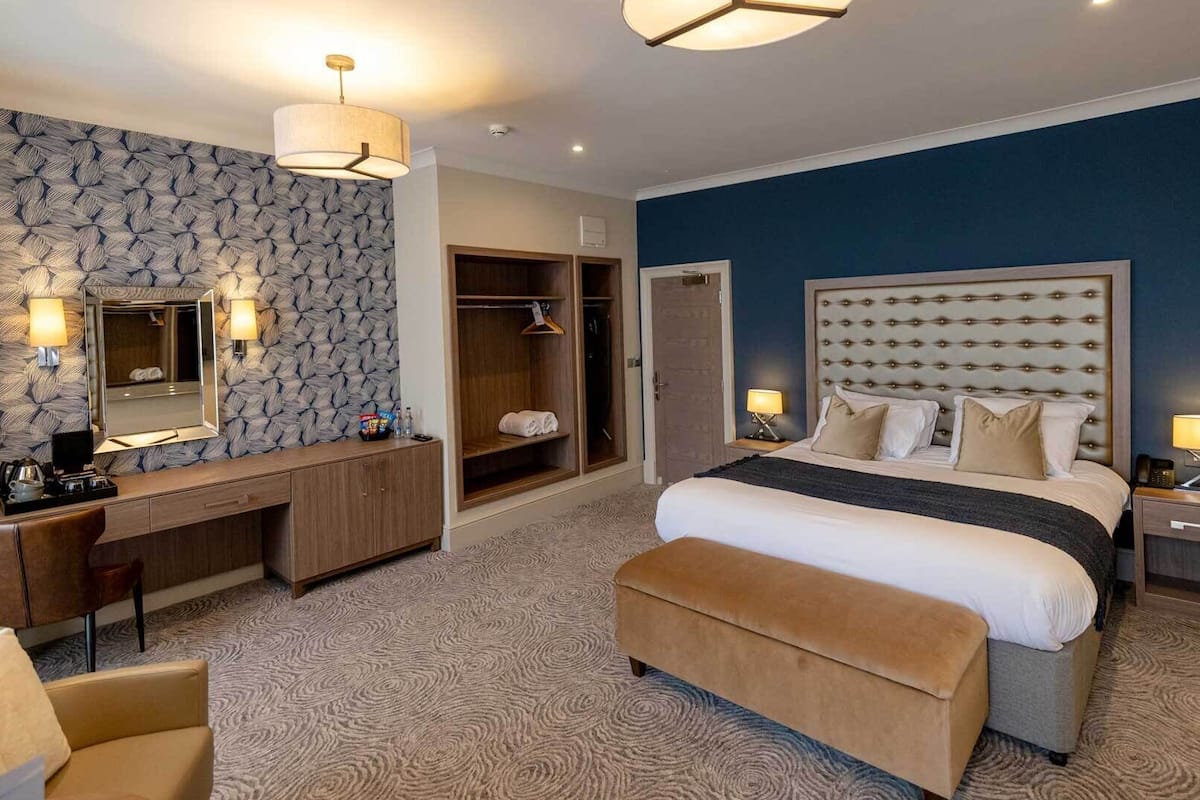 A modern hotel room with a large bed, a walk-in closet, a vanity with a mirror, and a seating area with a chair.