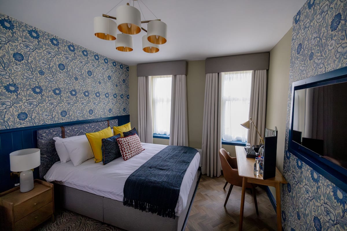 A hotel room with a bed, bedside table, lamp, desk with chair, and a television.