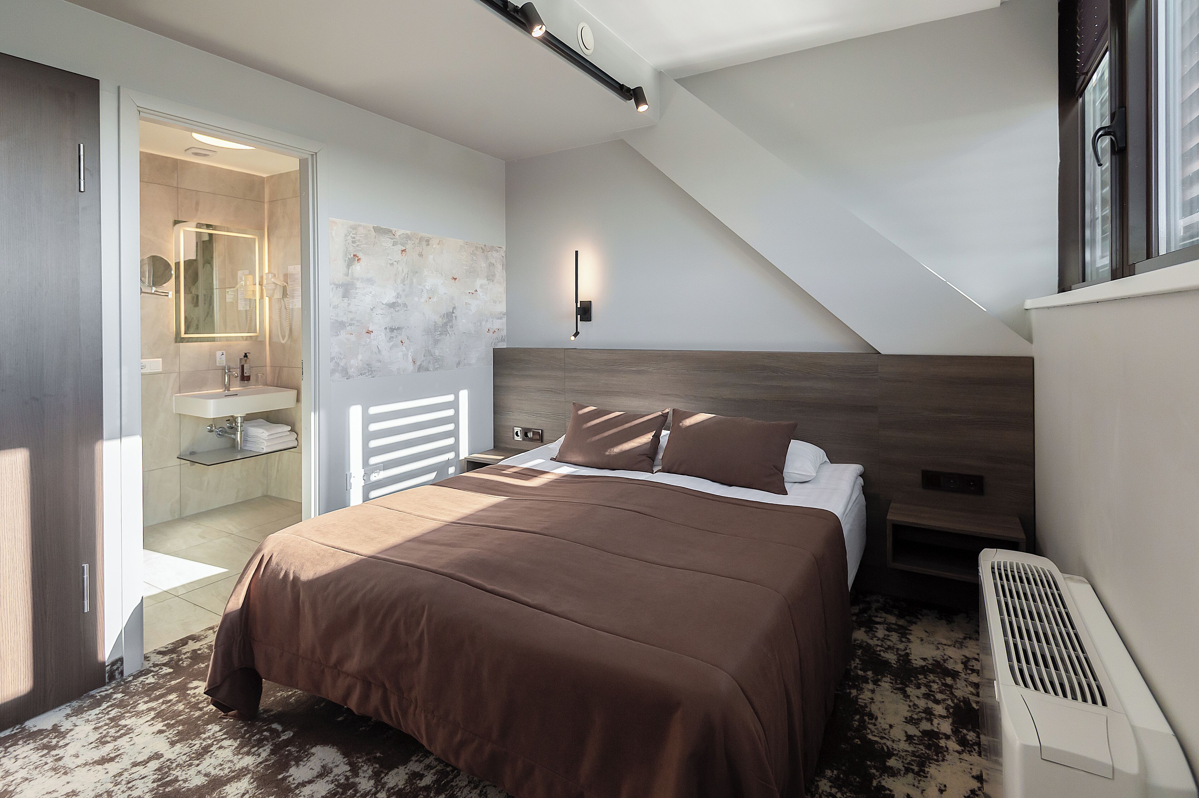 A bedroom with a large bed, a bedside table, a nightstand, a mirror, a wall-mounted light, and a window with blinds.