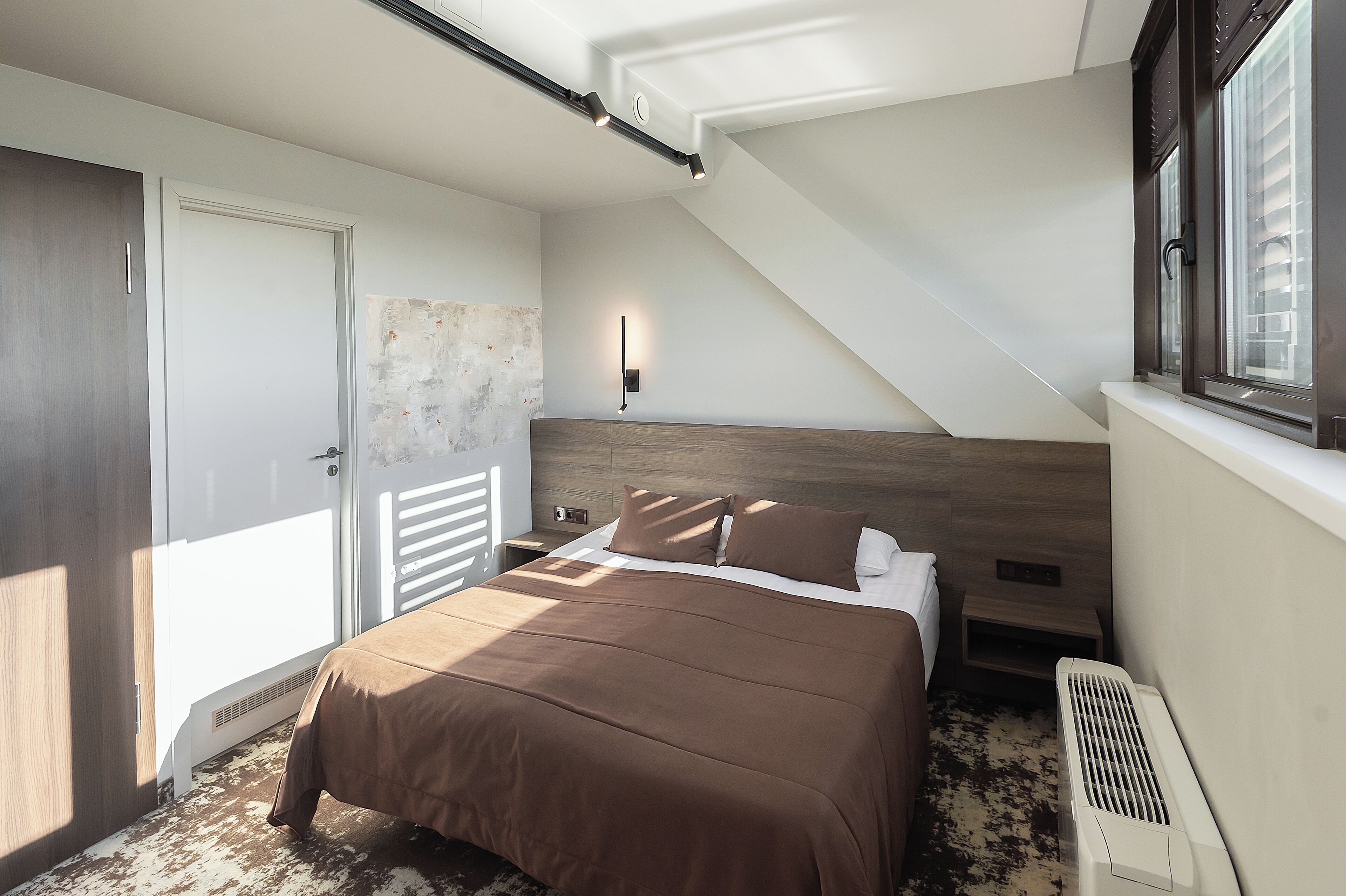 A bedroom with a bed, a nightstand, a wall-mounted lamp, a window with blinds, and a radiator.