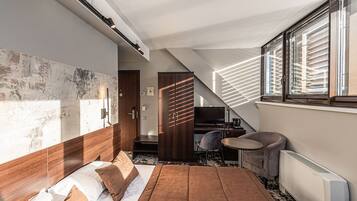 Ein modernes Hotelzimmer mit einem großen Bett, einem Schreibtisch und einem Stuhl. Es gibt ein Fenster mit Jalousien, eine Heizung und eine Tür zu einem anderen Raum.