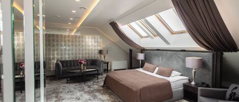 A modern hotel room with a large bed, a sofa, a small table, and a chair. There is a skylight above the bed and a view of the sky.
