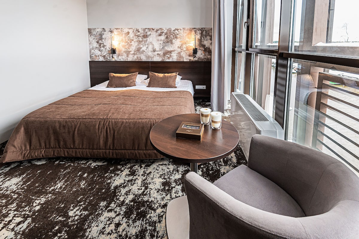 Ein Hotelzimmer mit einem großen Bett, einem runden Tisch mit einem Buch und zwei Gläsern, einem Sessel und Blick durch das Fenster.
