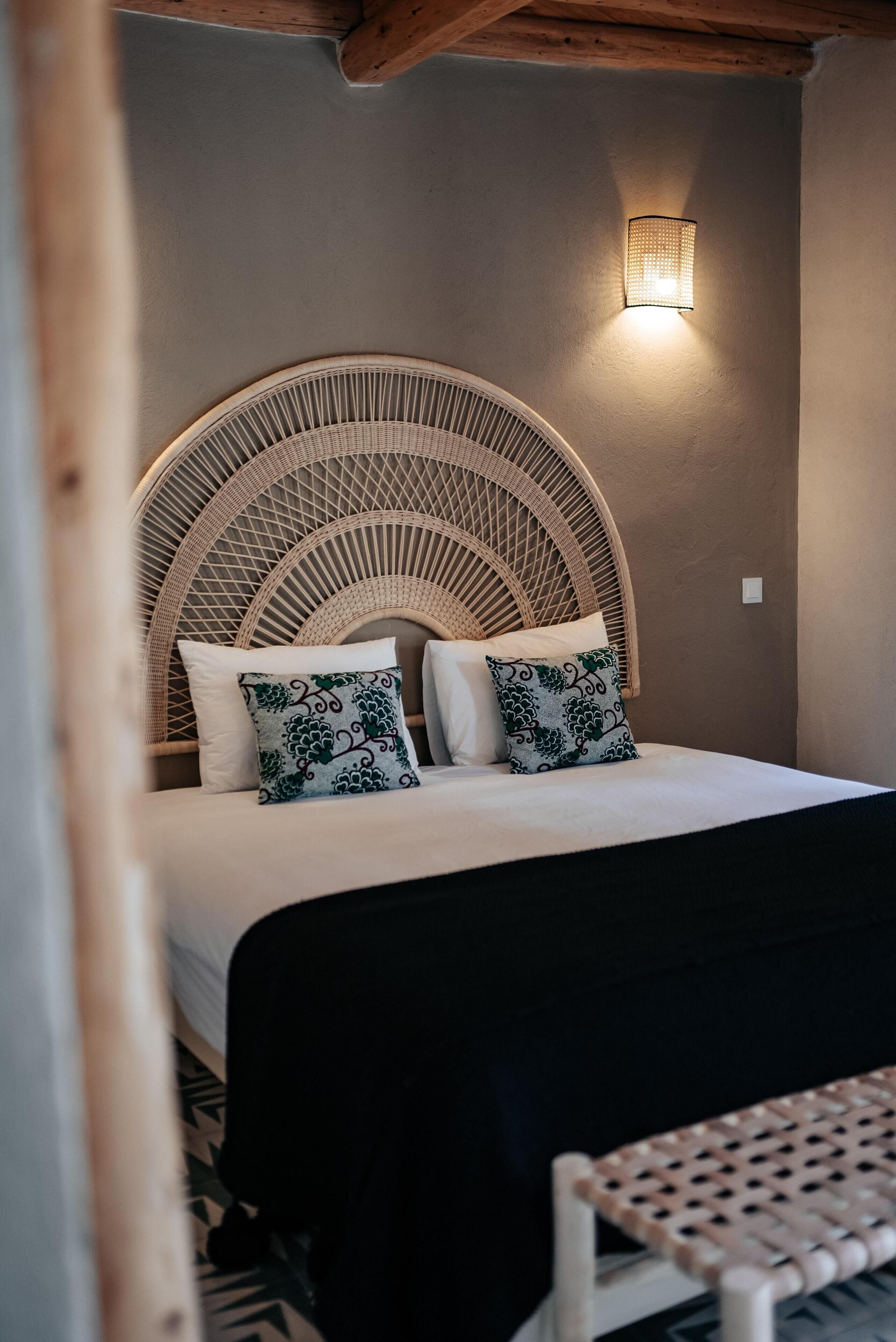 Una cama con cabecera trenzada, dos almohadas y una lámpara fijada a la pared.