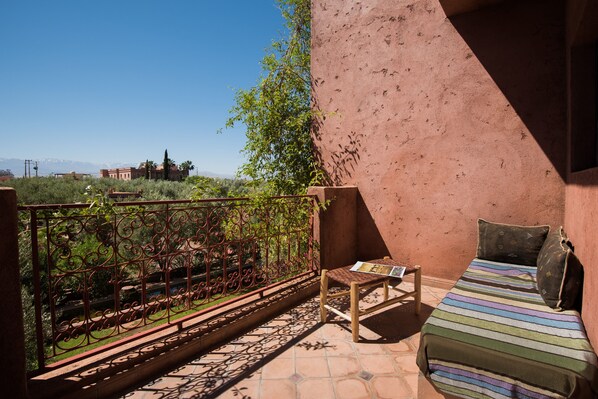 Un balcón con una cama, una mesita y vistas a un viñedo y edificios lejanos.