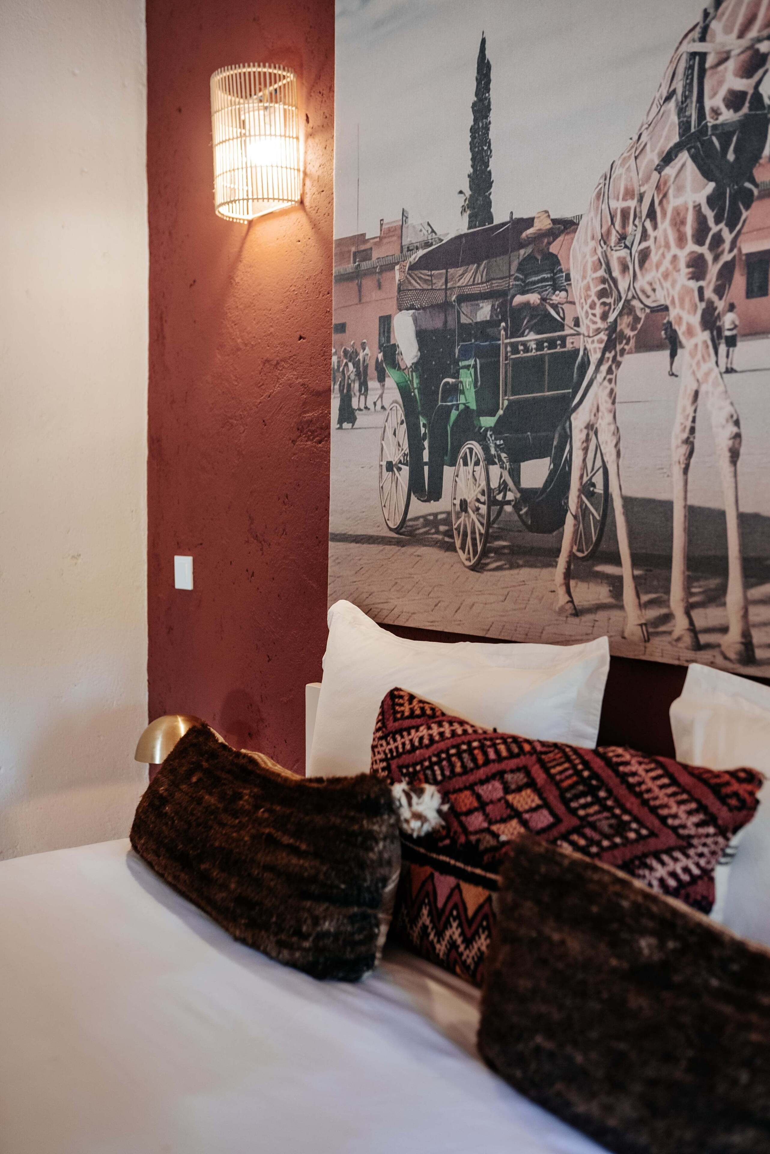 Ein Hotelzimmer mit Bett, Kissen, einer Wandleuchte und einem Wandgemälde mit Giraffe und Pferdekutsche.