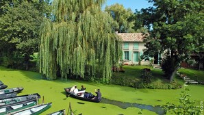 Une scène paisible au bord de la rivière, avec des bateaux amarrés le long de la rive, un saule pleureur et un bâtiment doté d’un toit en tuiles.