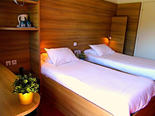 A hotel room with two single beds, a wooden headboard, a small table with a plant, and a shelf with a coffee maker.