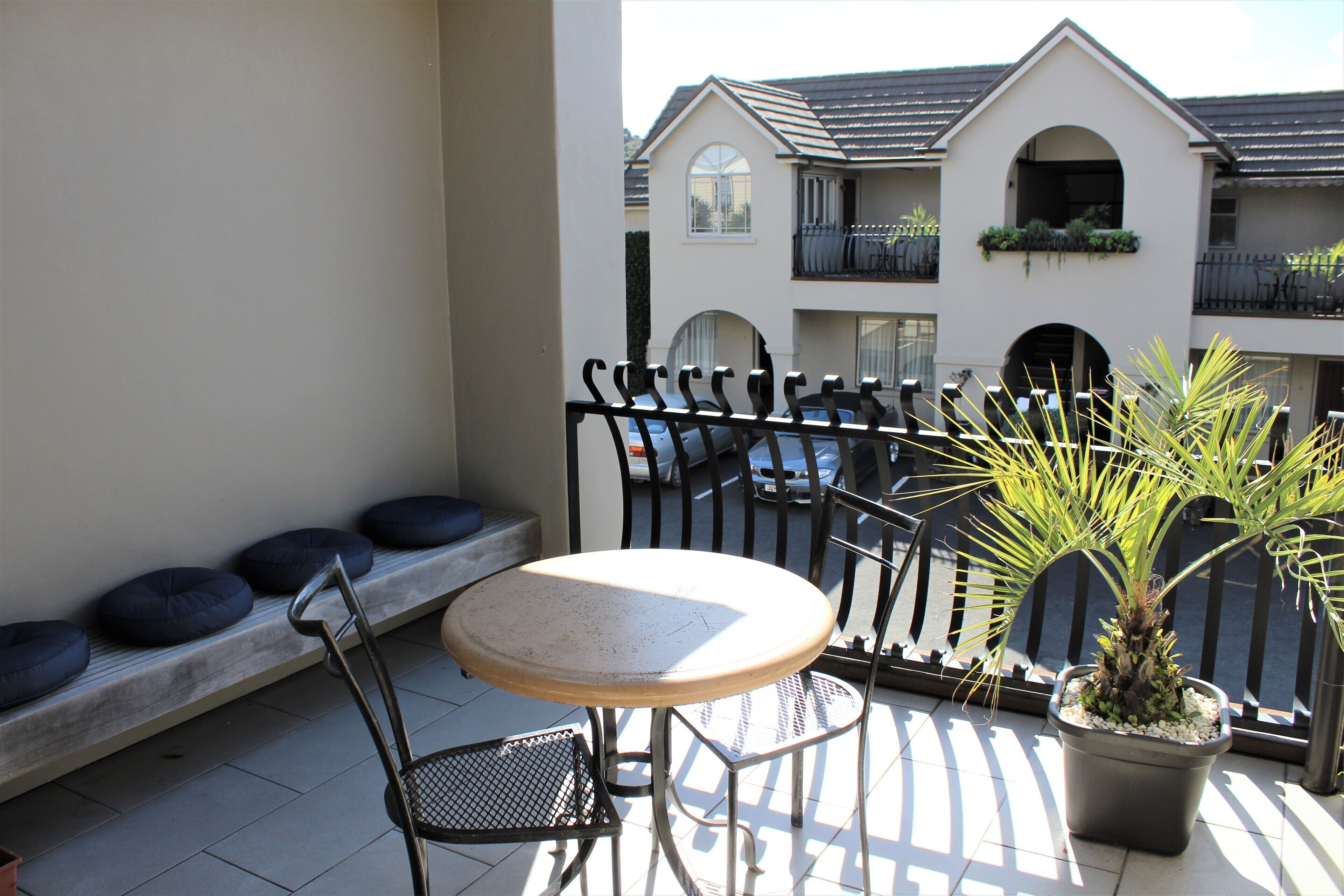 A balcony with a round table and two chairs, a potted plant, and a view of multi-story buildings in the background.
