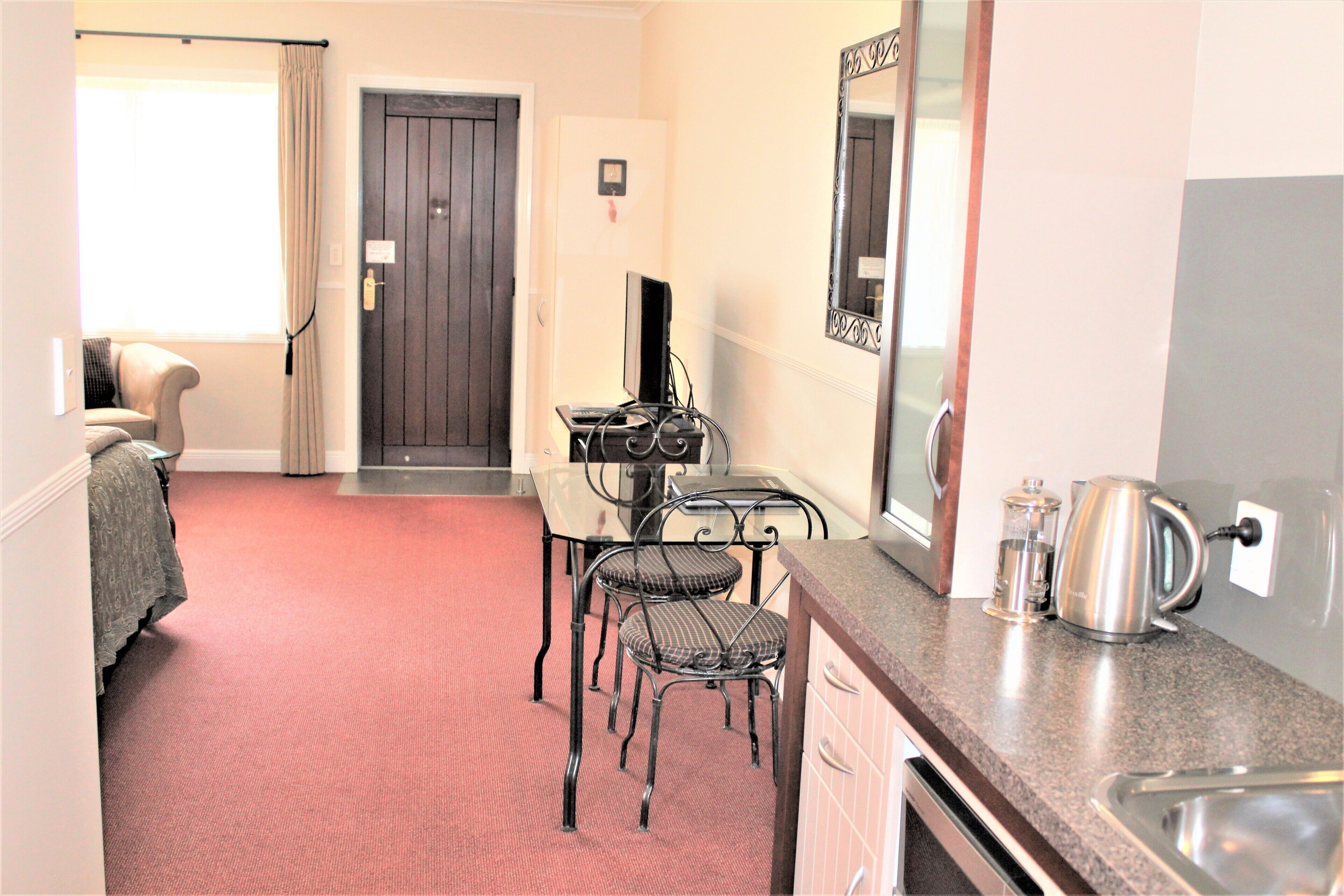 A compact hotel room with a kitchenette, a dining area with a table and chairs, a bed with a blanket, and a door leading to the outside.