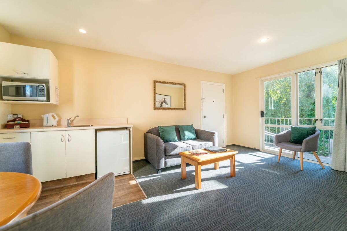 A compact living space with a kitchenette, a sofa, a coffee table, and a chair near a large window.
